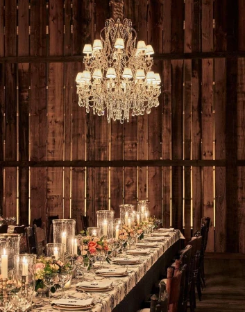 Crystal chandelier illuminates a long formal dining table set for an event in a barn interior with rustic wood walls and flooring.