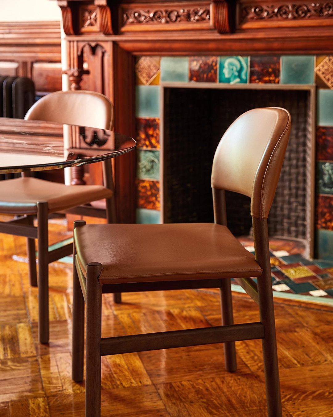 Interior design: Wooden chairs with tan leather seats are arranged around a glass table in a room with a decorative fireplace.