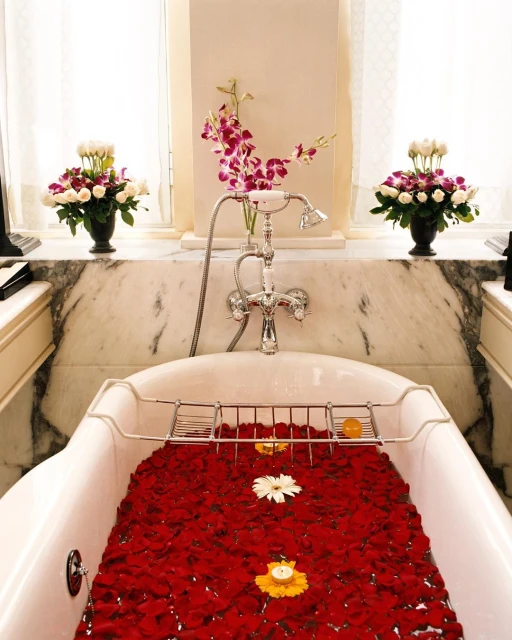 A bathtub is filled with red rose petals and illuminated by candlelight. Floral arrangements adorn the marble-topped surround.