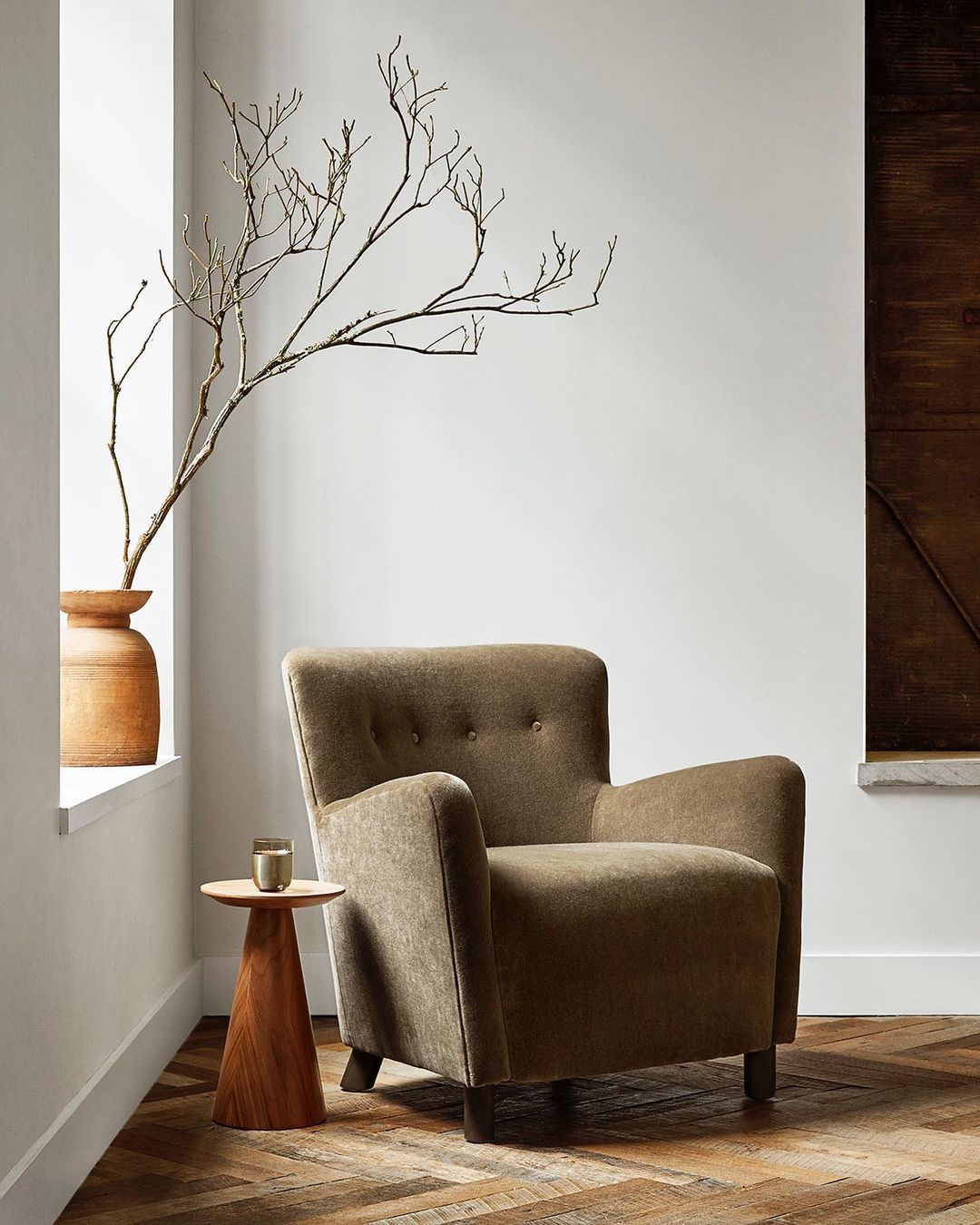 Interior view of a taupe armchair in a living room setting next to a side table, vase of branches, and a brown wood accent panel.