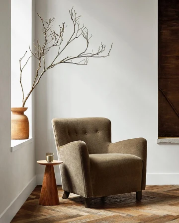 Interior view of a taupe armchair in a living room setting next to a side table, vase of branches, and a brown wood accent panel.