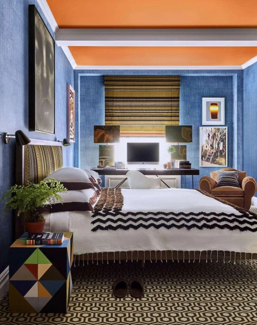 Interior of a bedroom with a bed, patterned rug, a colorful side table with books and plant, with desk and chair in the background.