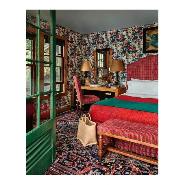 A bedroom features floral wallpaper, a desk, a bed with red and green covers, and a woven bag on a patterned rug, inviting and eclectic.