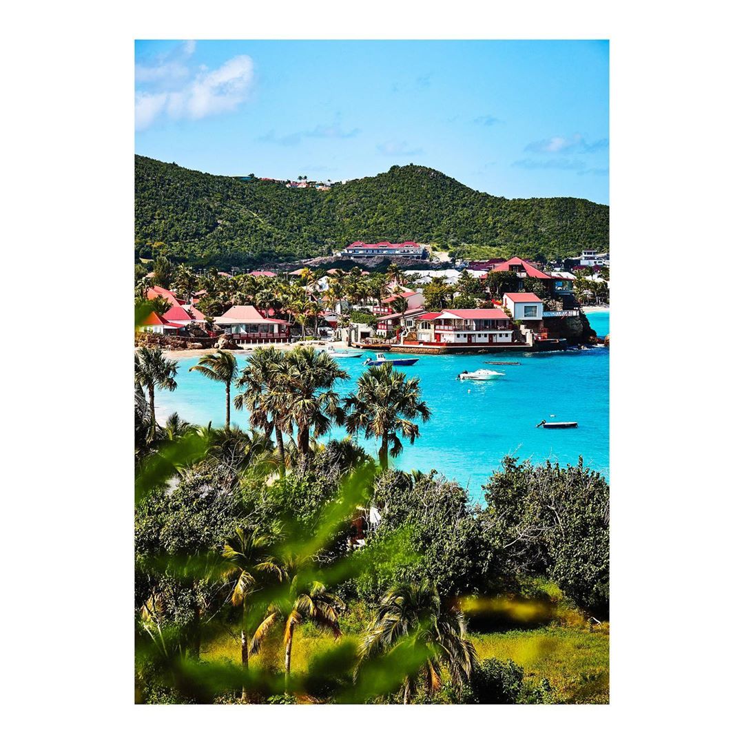 Coastal view of tropical island with palm trees, turquoise water, boats, and hillside homes against a bright blue sky on a sunny day.