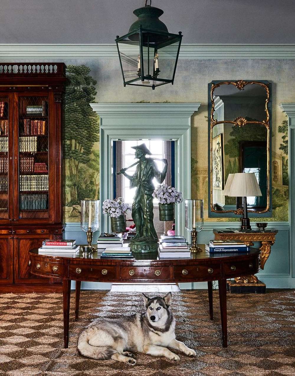 Living room interior featuring a husky dog lying on a patterned rug, dark wood table with books and flowers, a statue, and wallpaper.