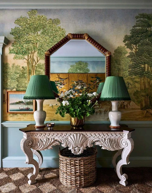 Interior view of a console table with lamps and flowers, set against a backdrop of landscape-themed wallpaper and a decorative mirror.