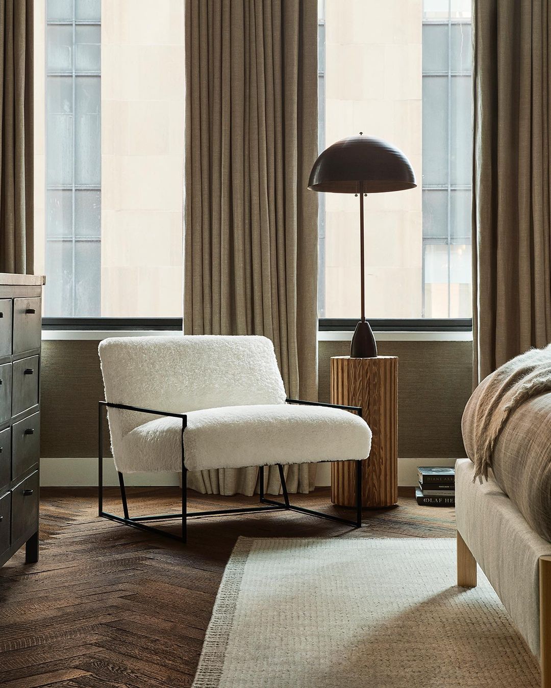 Interior photograph of a modern living room with a white armchair, wooden side table with a lamp, dark wood floors and large windows.