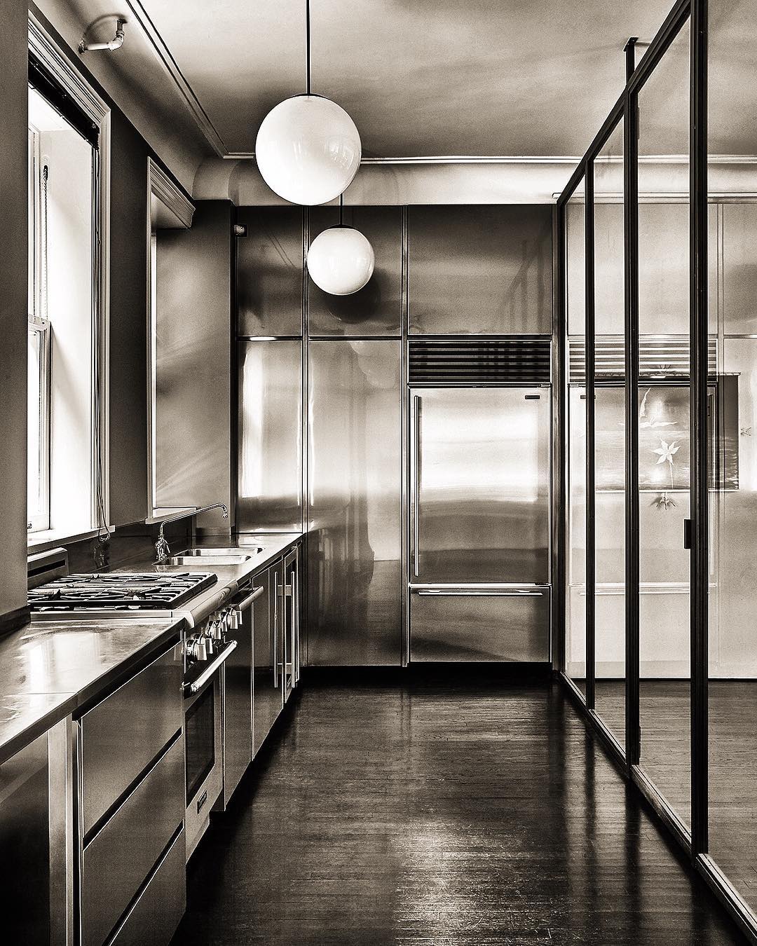 Modern stainless steel kitchen interior with dark wood floors, stainless steel appliances, globe pendant lights, and framed glass panels.