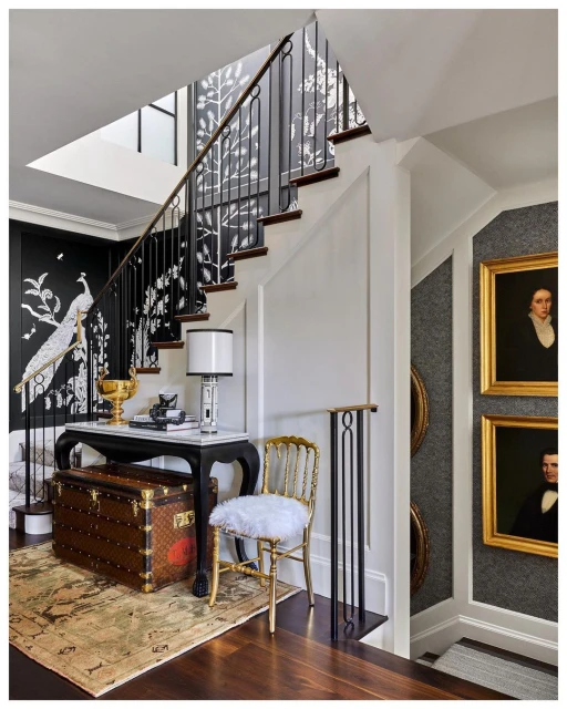 Elegant entryway featuring a staircase with iron railing, a console table with lamp, framed artwork, and a Louis Vuitton trunk.