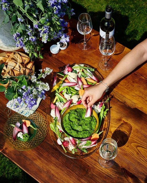 Overhead shot of a wooden table with a vegetable platter, dip, wine, glasses, flowers, and a woman's hand dipping a vegetable.