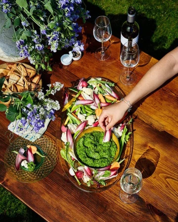 Overhead shot of a wooden table with a vegetable platter, dip, wine, glasses, flowers, and a woman's hand dipping a vegetable.
