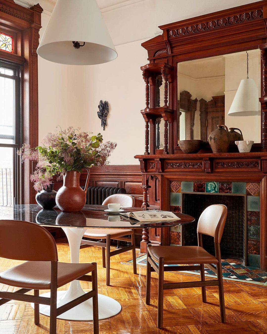 Interior dining room with a round marble table, brown leather chairs, a wooden fireplace mantle, and a white pendant light overhead.