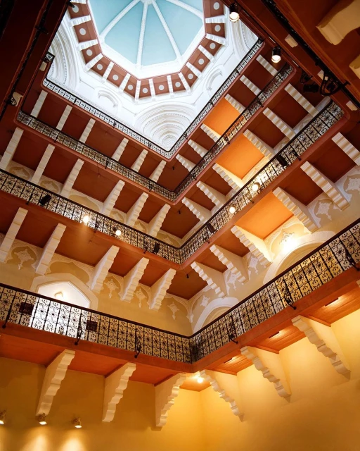 Indoor architectural shot looking up at a building atrium with multi-tiered balconies and a skylight dome in a square symmetrical format.