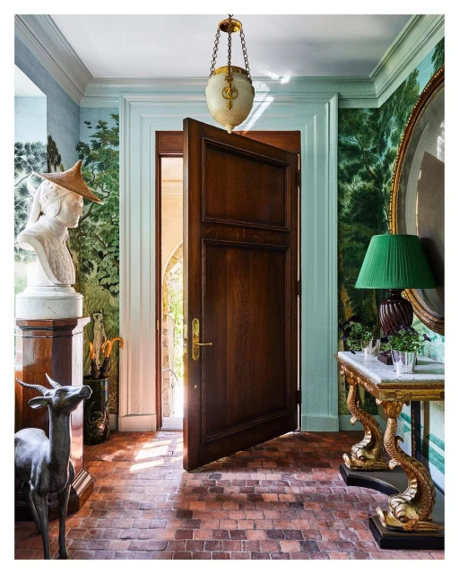 Elegant interior entryway with open wooden door, marble-topped console table, green lampshade, statue, and intricate forest-themed wallpaper.