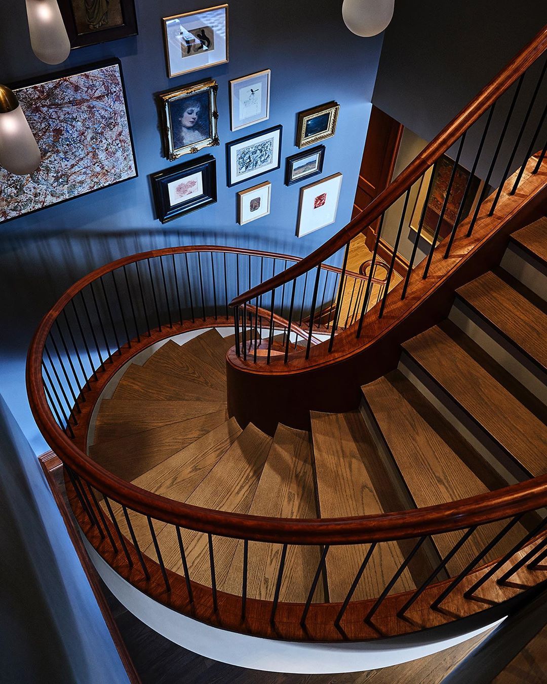 A high-angle shot captures a winding wooden staircase with a dark wood railing in an interior setting, artwork decorates the walls.