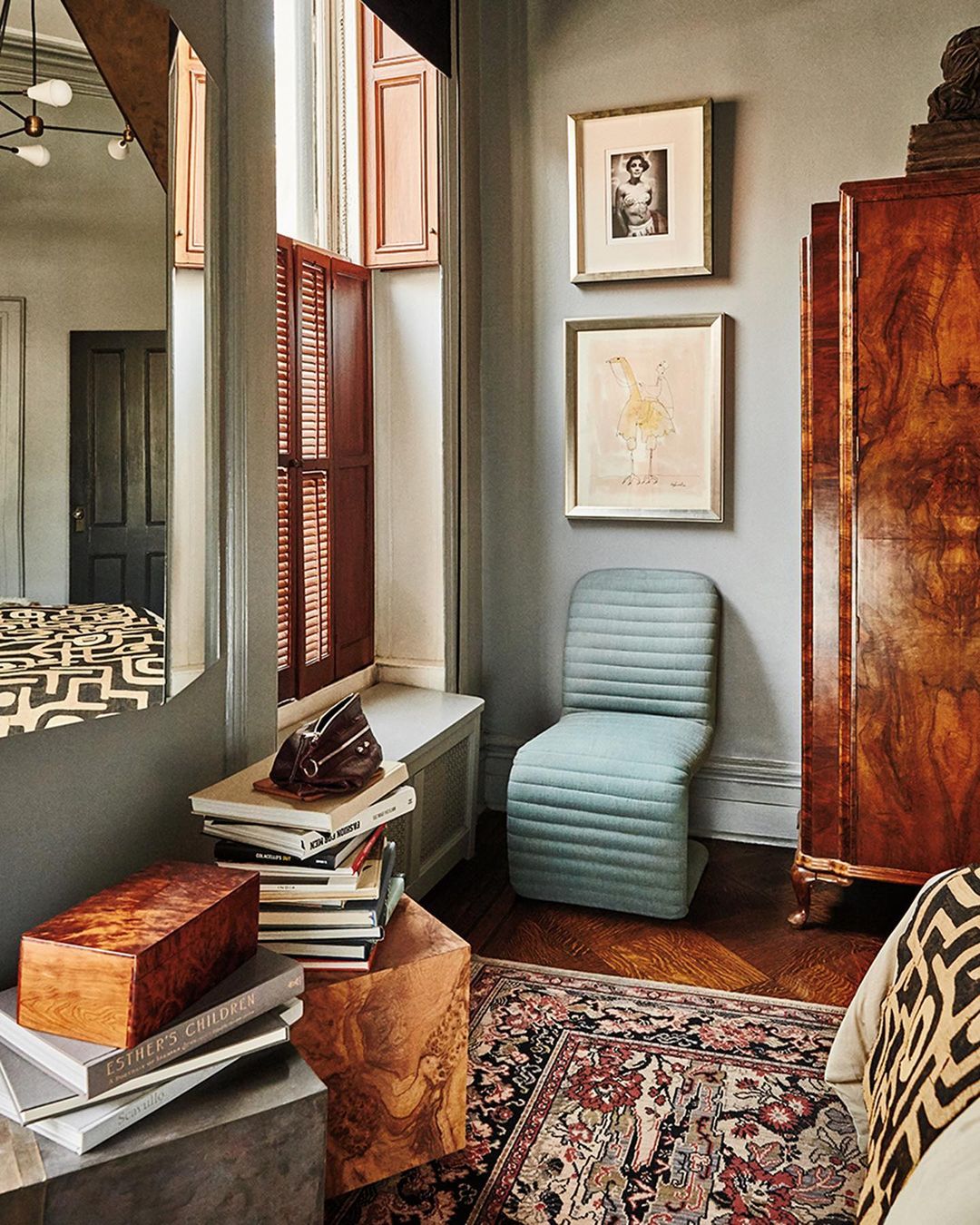 Eclectic interior with a blue chair, antique cabinet, patterned rug, and books. Natural light filters through shuttered windows.