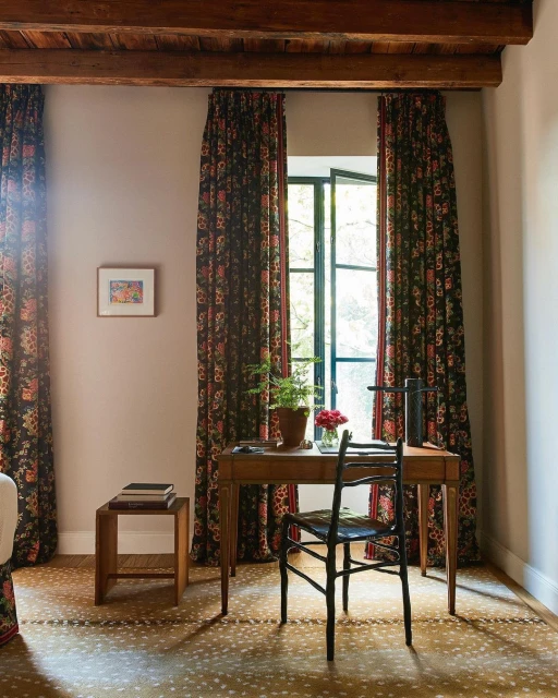 Home office interior featuring a wooden desk with flowers, floral curtains, wood-beamed ceiling, and patterned carpet.