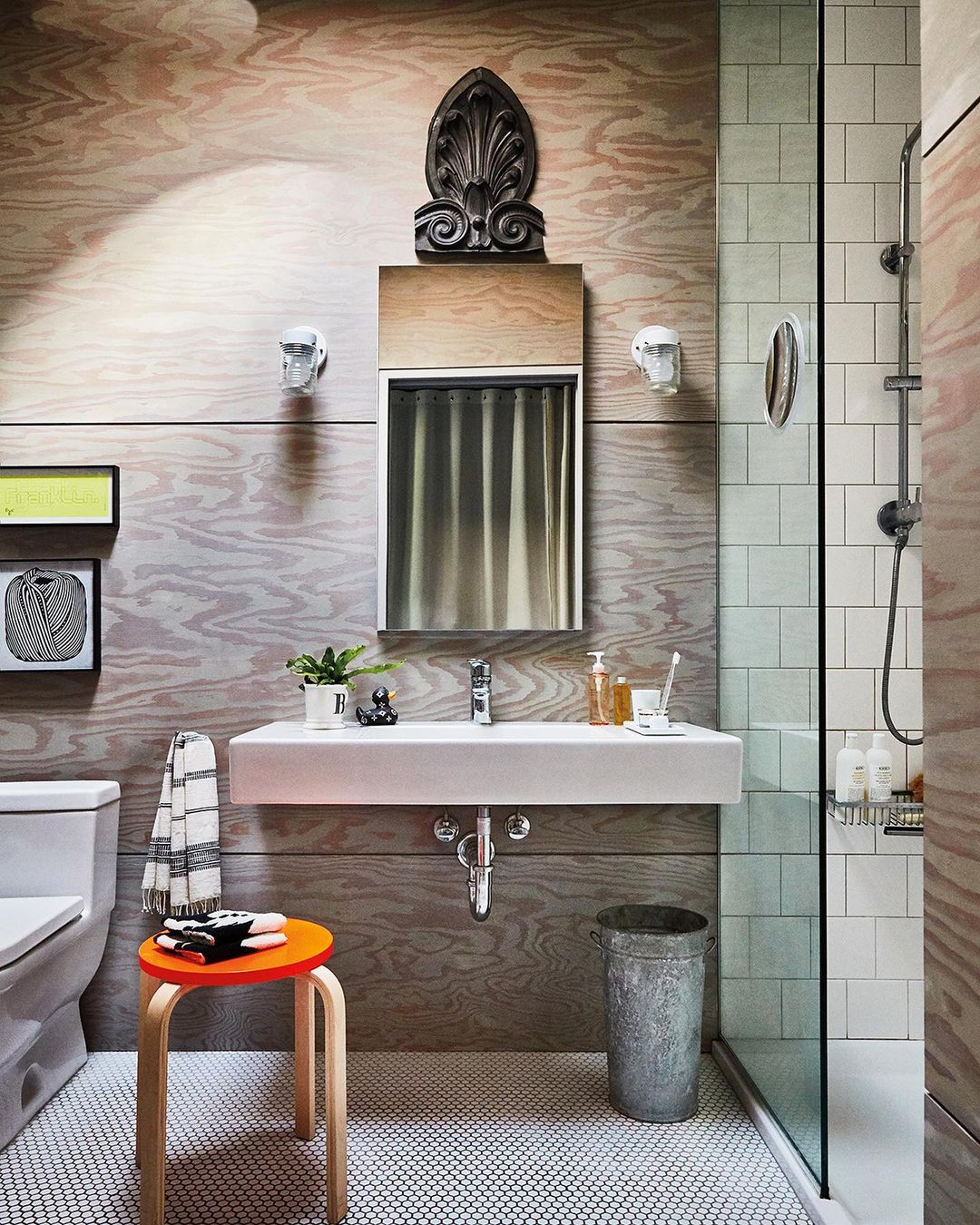 Bathroom interior featuring a modern vanity, a toilet, an orange stool, and a glass-enclosed shower with white tiled walls and wooden accents.