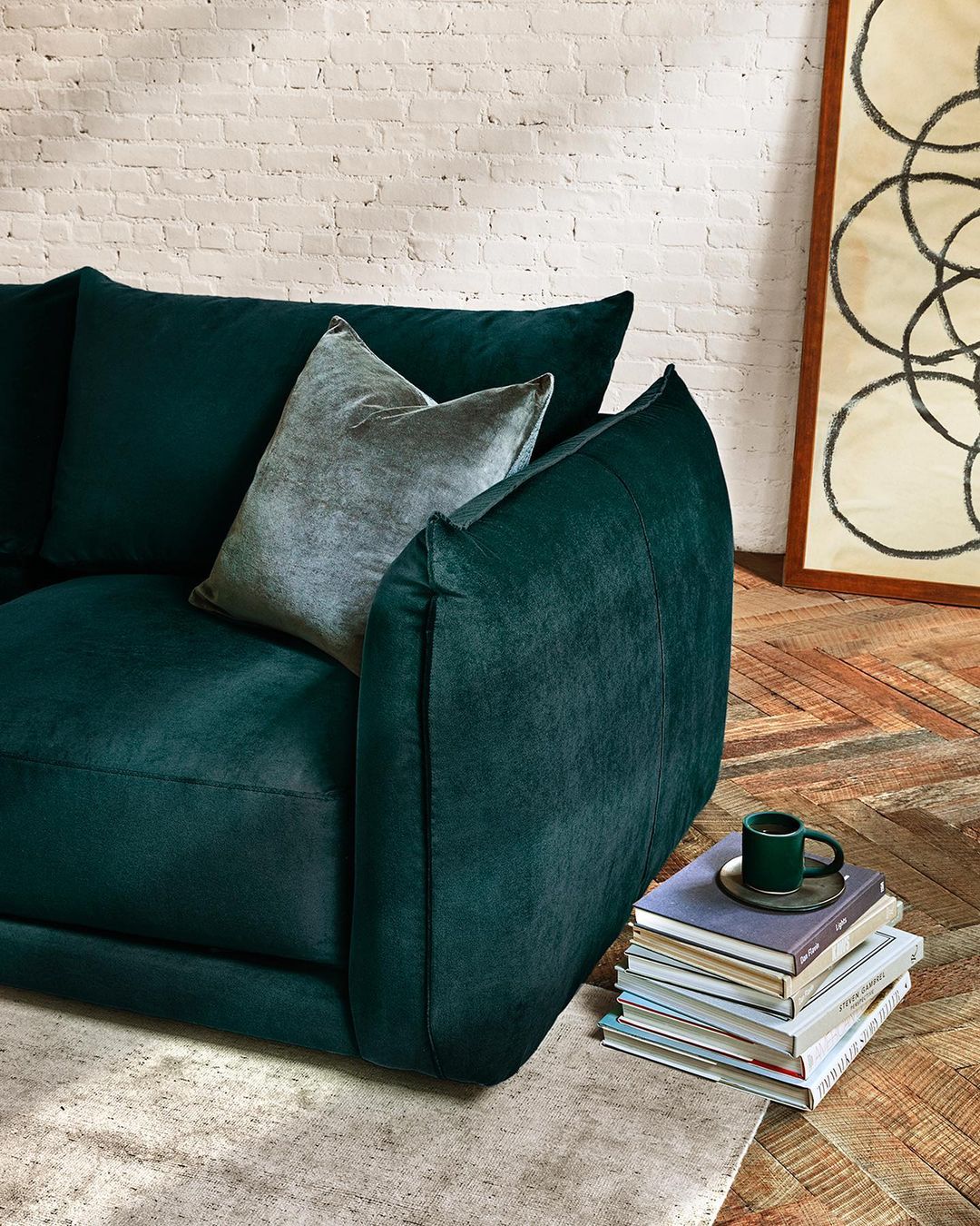 Living room interior featuring a dark green velvet sofa with a throw pillow, a stack of books, and decorative art, indoors.