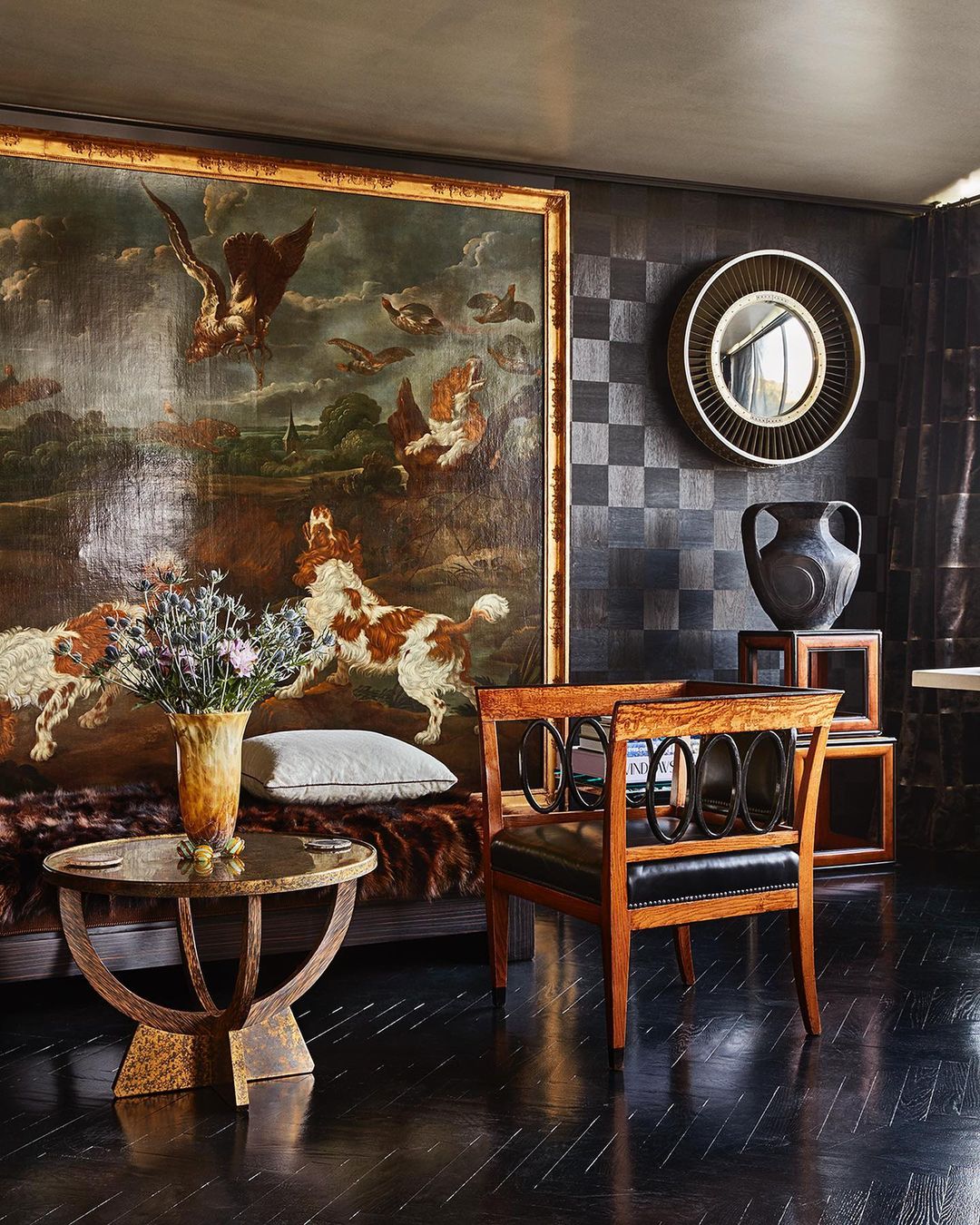 Interior of a living room featuring a large painting, a vase of flowers, a round table, a decorative mirror, and dark wooden floors.