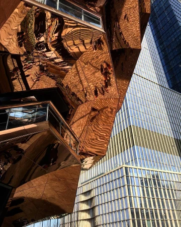 Low-angle shot of the Vessel sculpture with skyscraper backdrop in Hudson Yards, NYC, showcasing architecture and modern cityscape.
