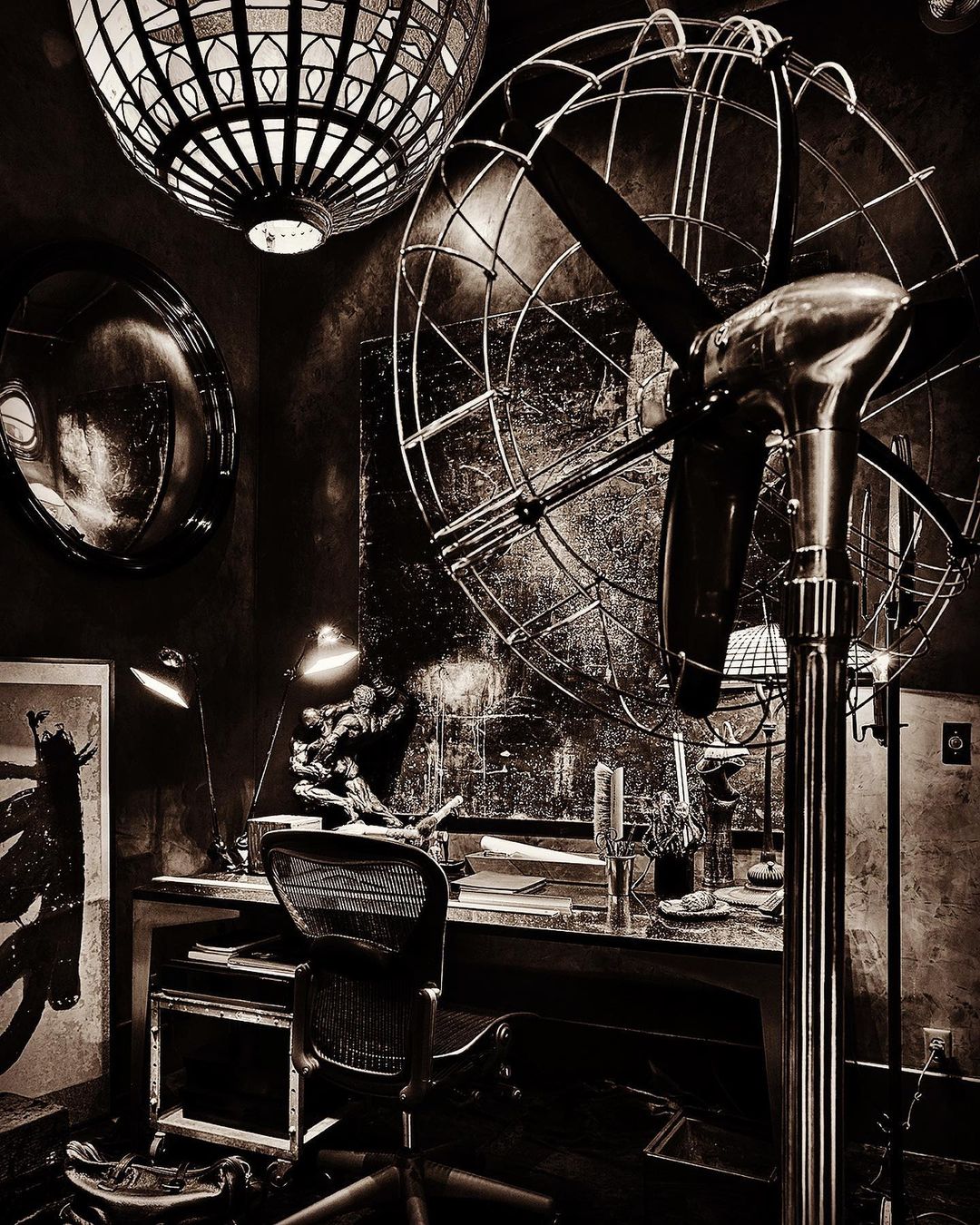 Indoor photo of a workspace featuring a standing fan, desk with objects, lamp, chair, and decorative lighting, captured with brown color grading.