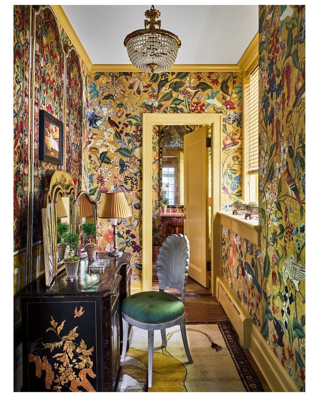 Ornate interior hallway with floral wallpaper, antique furniture, chandelier, and an open doorway leading to another room.