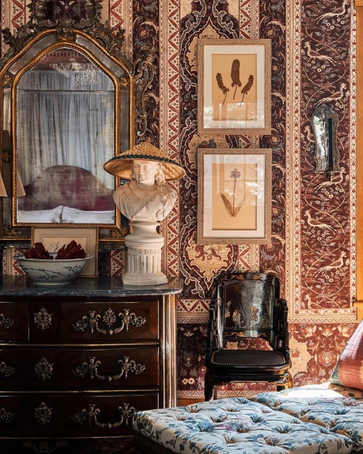 Ornate interior featuring a bust with a straw hat, a framed mirror reflecting a bed, framed botanical prints, and dark wood furniture.
