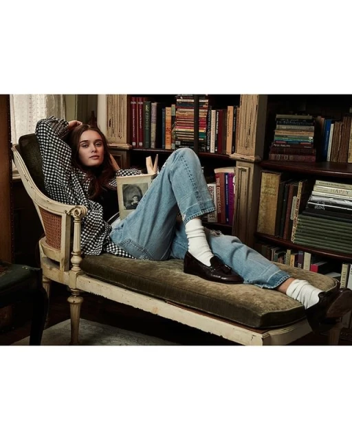 Young woman relaxes in an antique chair, casually reading a book in a home library setting with shelves filled with books and natural light.
