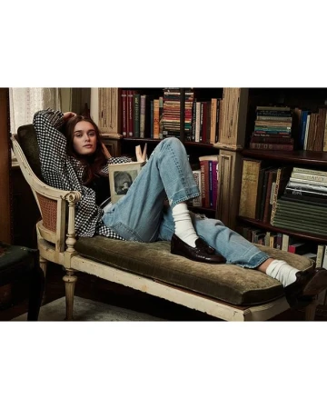 Young woman relaxes in an antique chair, casually reading a book in a home library setting with shelves filled with books and natural light.