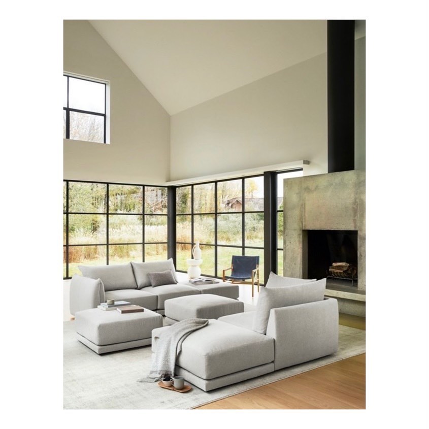 Minimalist living room interior featuring a grey modular sofa, large windows, a concrete fireplace, and natural light, creating a serene atmosphere.