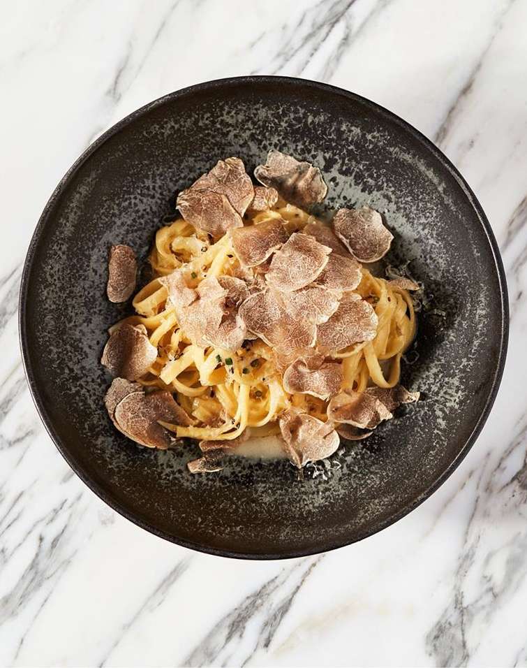 Overhead shot of tagliatelle pasta with shaved black truffle on a black plate, set on a white marble surface.