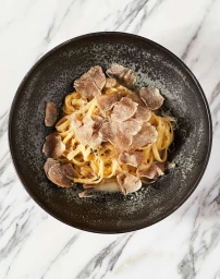 Bowl of fresh pasta topped with sliced truffles served in a dark ceramic bowl on a marble tabletop