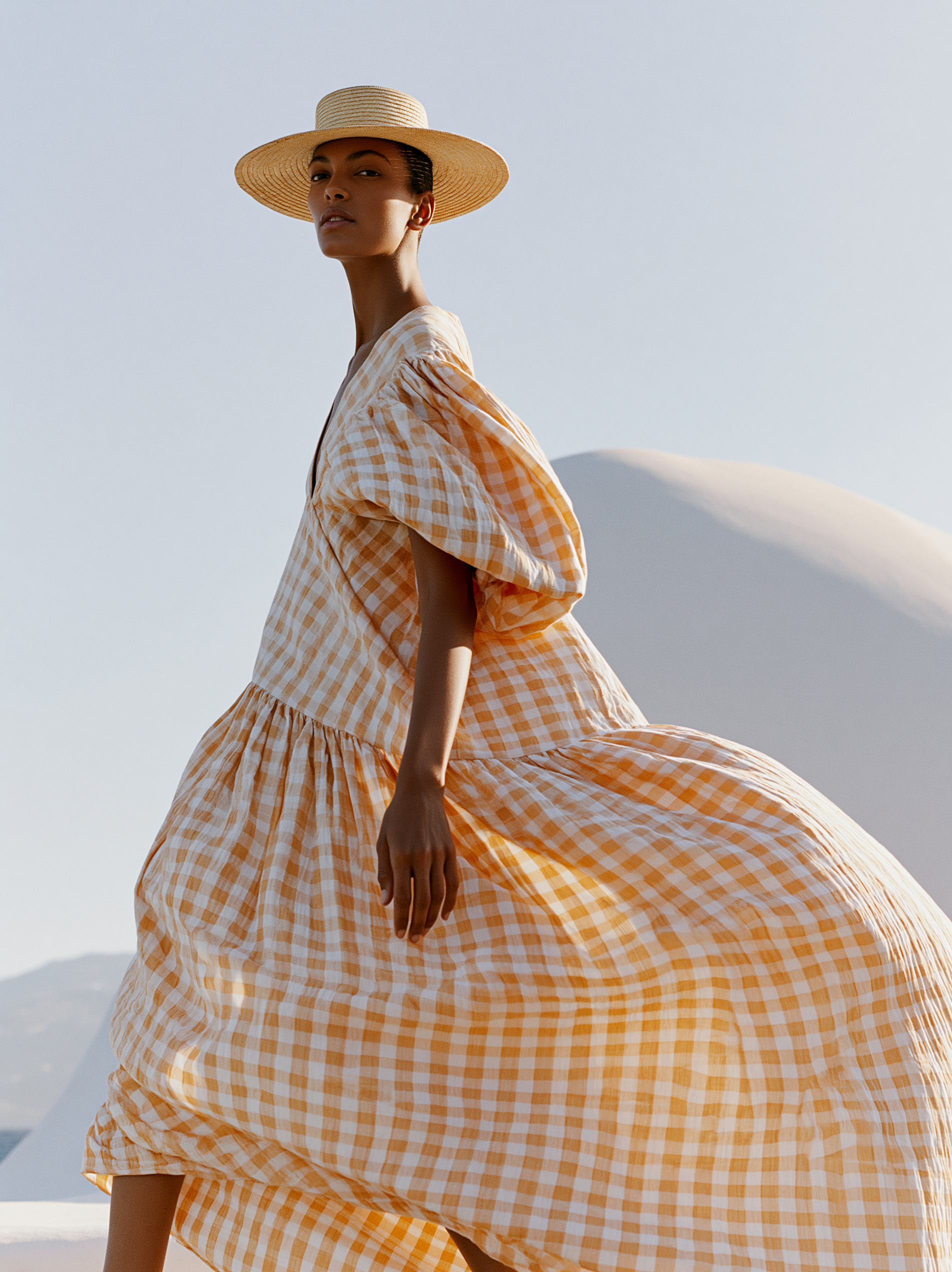 A woman wearing a straw hat and a light orange and white gingham midi dress stands outdoors against a bright sky and soft dunes.