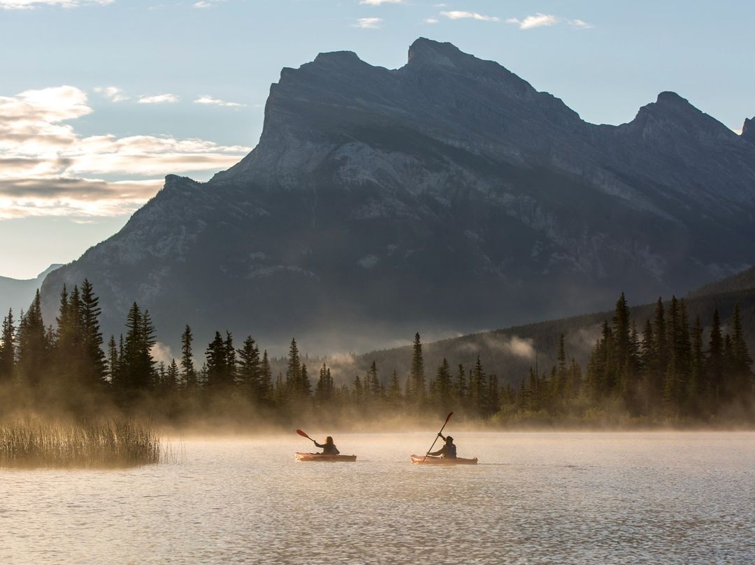 Two people kayaking on a lake with fog and tall pine trees in the early morning with a view of mountains in the background.