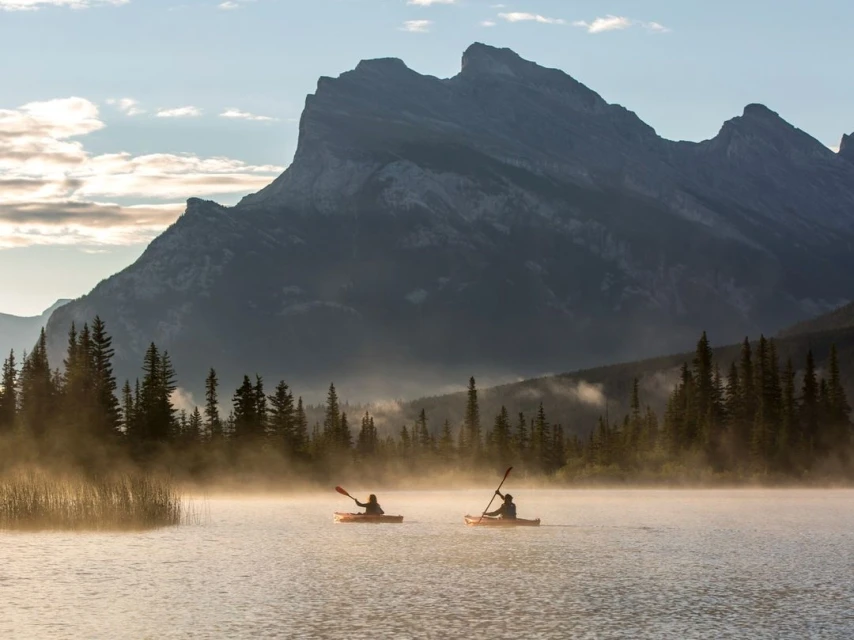 Two people kayaking on a lake with fog and tall pine trees in the early morning with a view of mountains in the background.