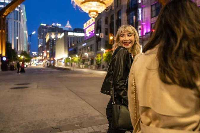 Two women walking on a city street at night, with one smiling over her shoulder wearing a black leather jacket.