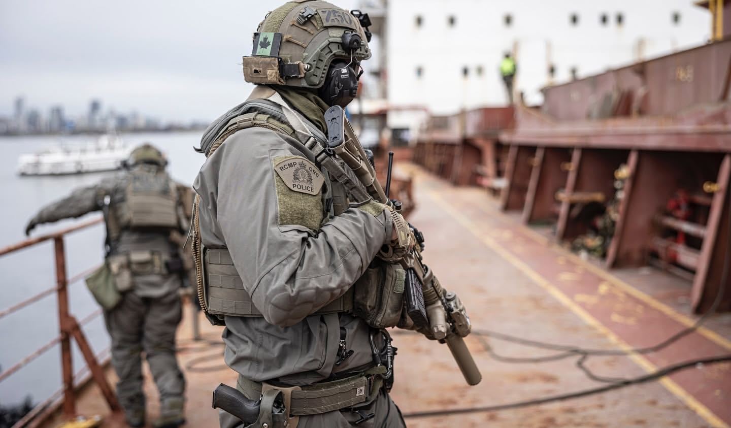 Two RCMP officers in tactical gear stand on a ship, one holding a rifle, with a city skyline visible in the background against a cloudy sky.
