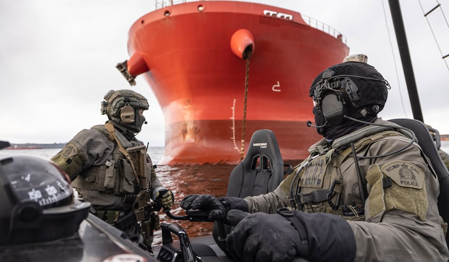 Two RCMP officers wearing tactical gear in a rigid hull inflatable boat (RHIB) with a large red cargo ship in the background.