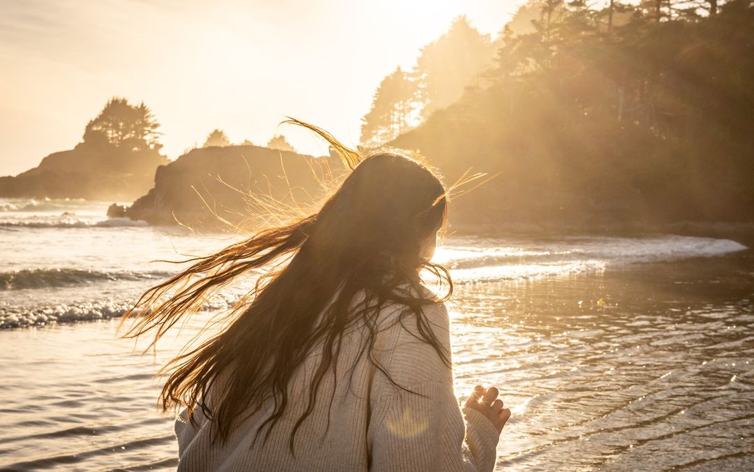 Woman with long brown hair stands on a beach at sunset, back to camera, wearing a white sweater, watching ocean waves roll in.