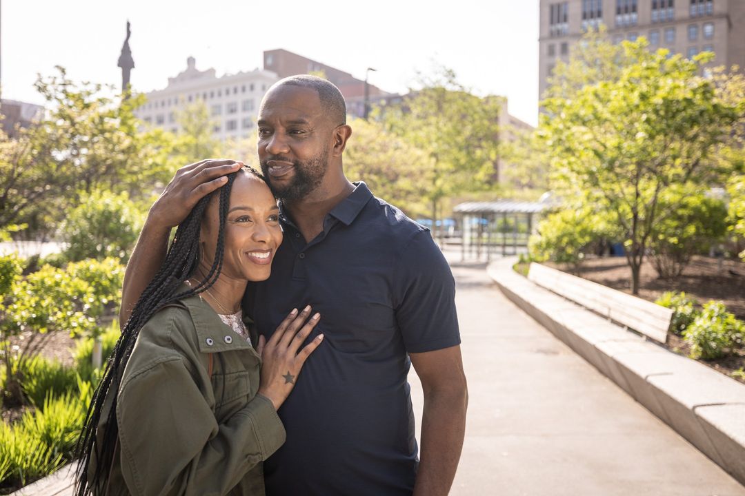 A smiling African American couple in a park setting embrace with natural sunlight, creating a warm, inviting atmosphere with city buildings.