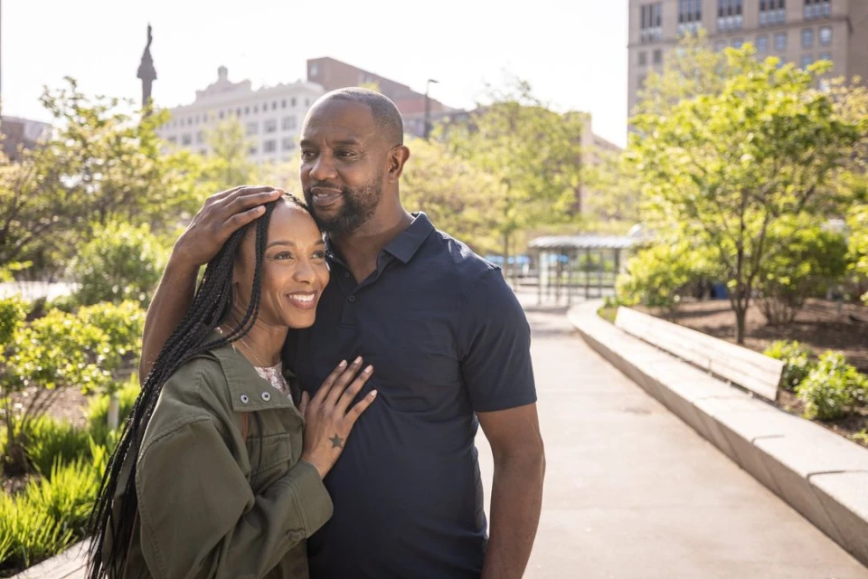 A smiling African American couple in a park setting embrace with natural sunlight, creating a warm, inviting atmosphere with city buildings.
