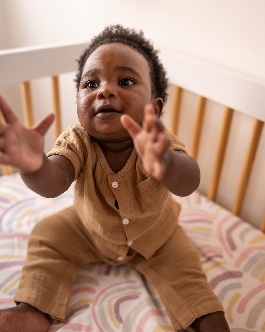 Close-up of an adorable African-American baby wearing a brown outfit, sitting in a white crib with rainbow-patterned bedding, looking upwards.