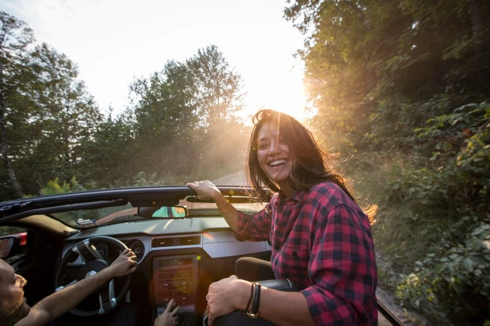 A woman smiles from a convertible as the sun shines through a forest. A man drives the red car on a sunlit rural road.