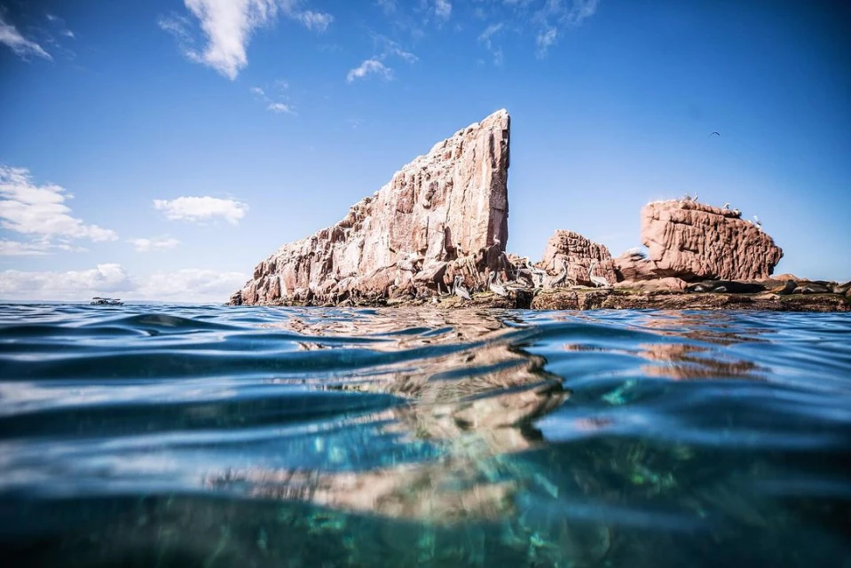 Large rock formations emerge from the ocean beneath a blue sky with scattered clouds, water surface showing sea life below.