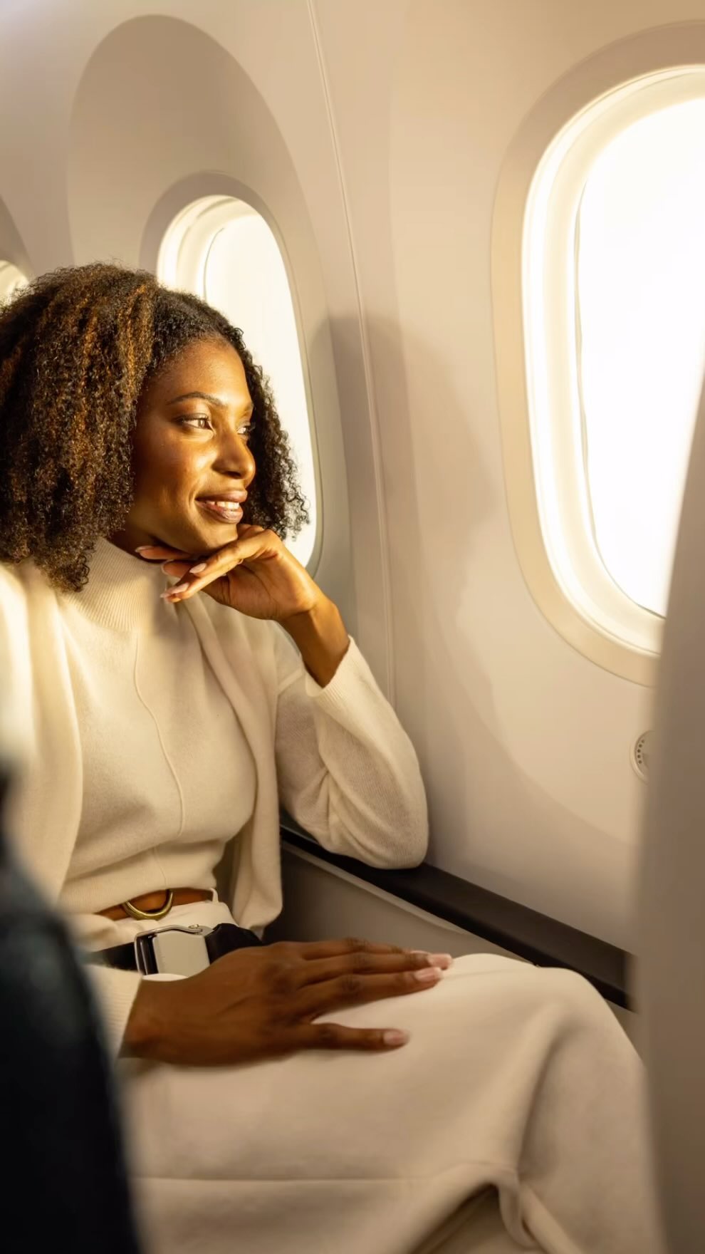 Woman in an airplane seat looks out the window, dressed in white, with natural light and a focus on travel and lifestyle.