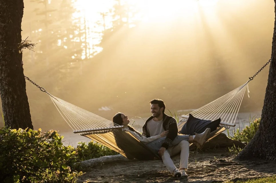 A couple relaxes together in a hammock strung between trees in a forest at sunset. Golden sunlight filters through the branches.