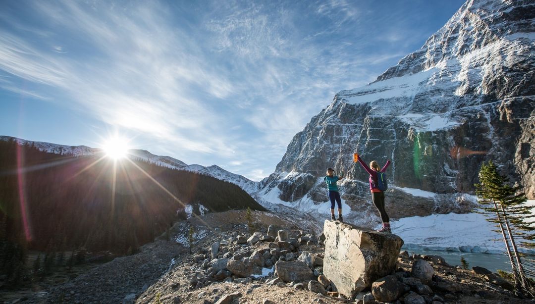 Two hikers celebrate atop a rock in a scenic mountain landscape under a bright sun, showcasing adventure and natural beauty.