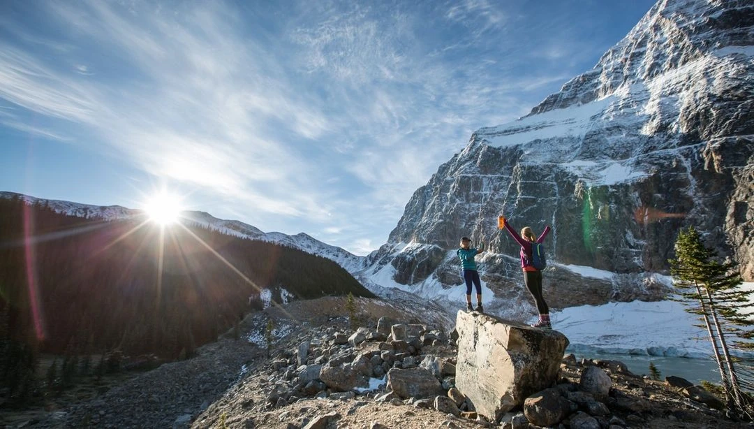 Two hikers celebrate atop a rock in a scenic mountain landscape under a bright sun, showcasing adventure and natural beauty.