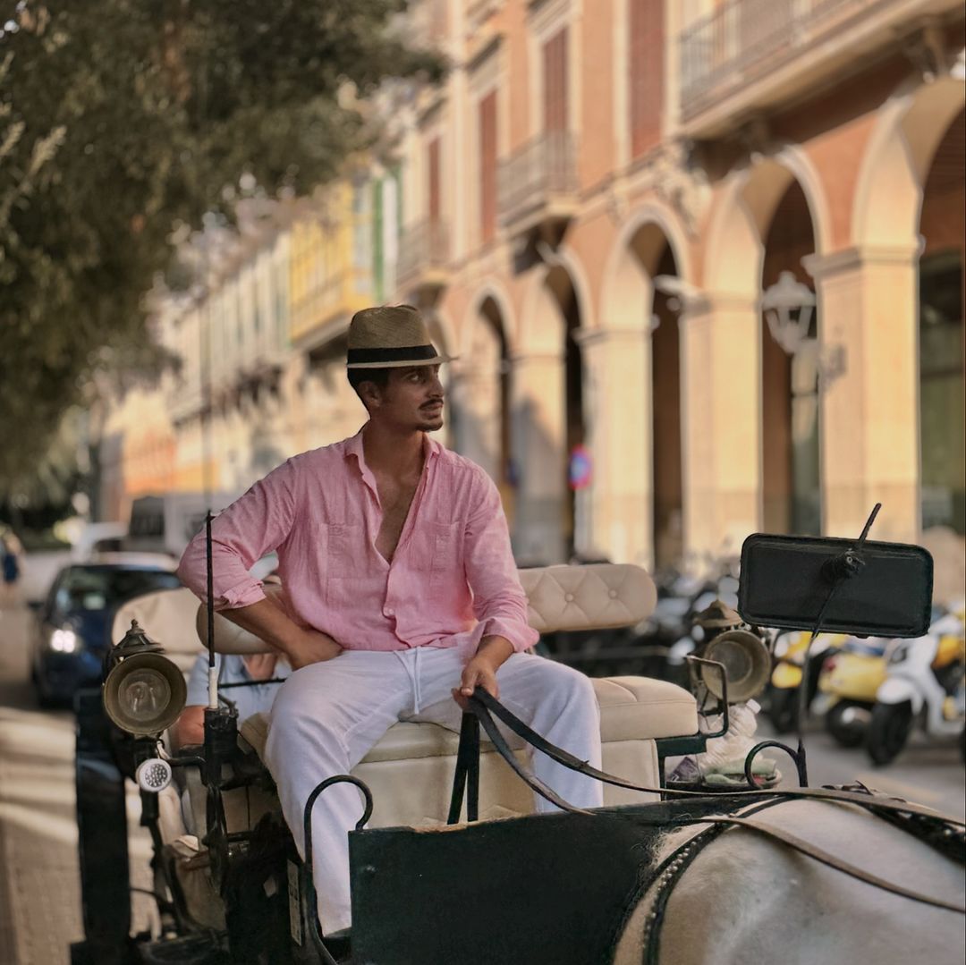 Adult male in a hat sits on a horse-drawn carriage on a European city street with old buildings and parked scooters on a sunny day.